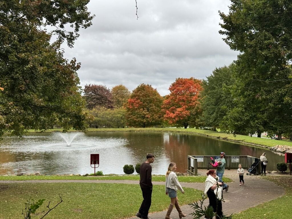 Families stroll past a pond with a fountain and orange maple trees at White House Fruit Farm